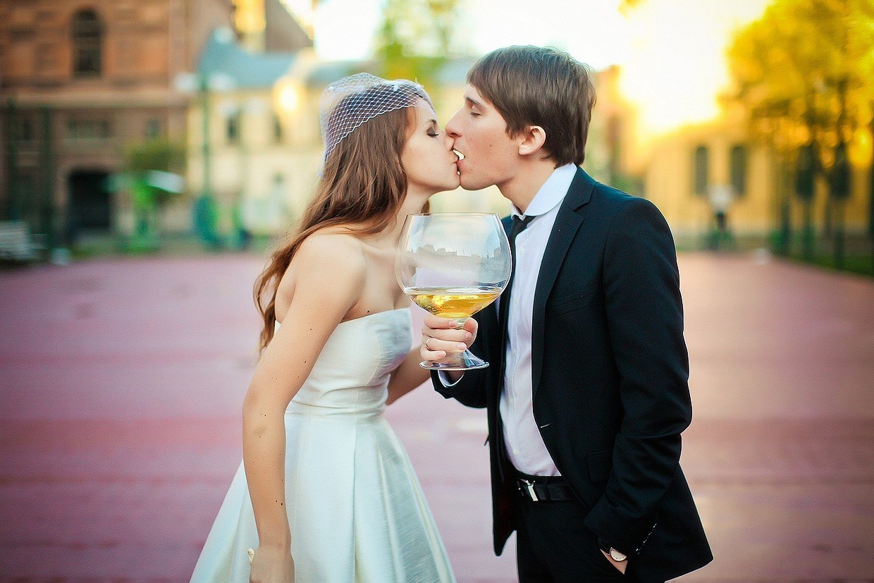 невеста в маленькой шляпке держит в руках огромный бокал для вина и целует жениха