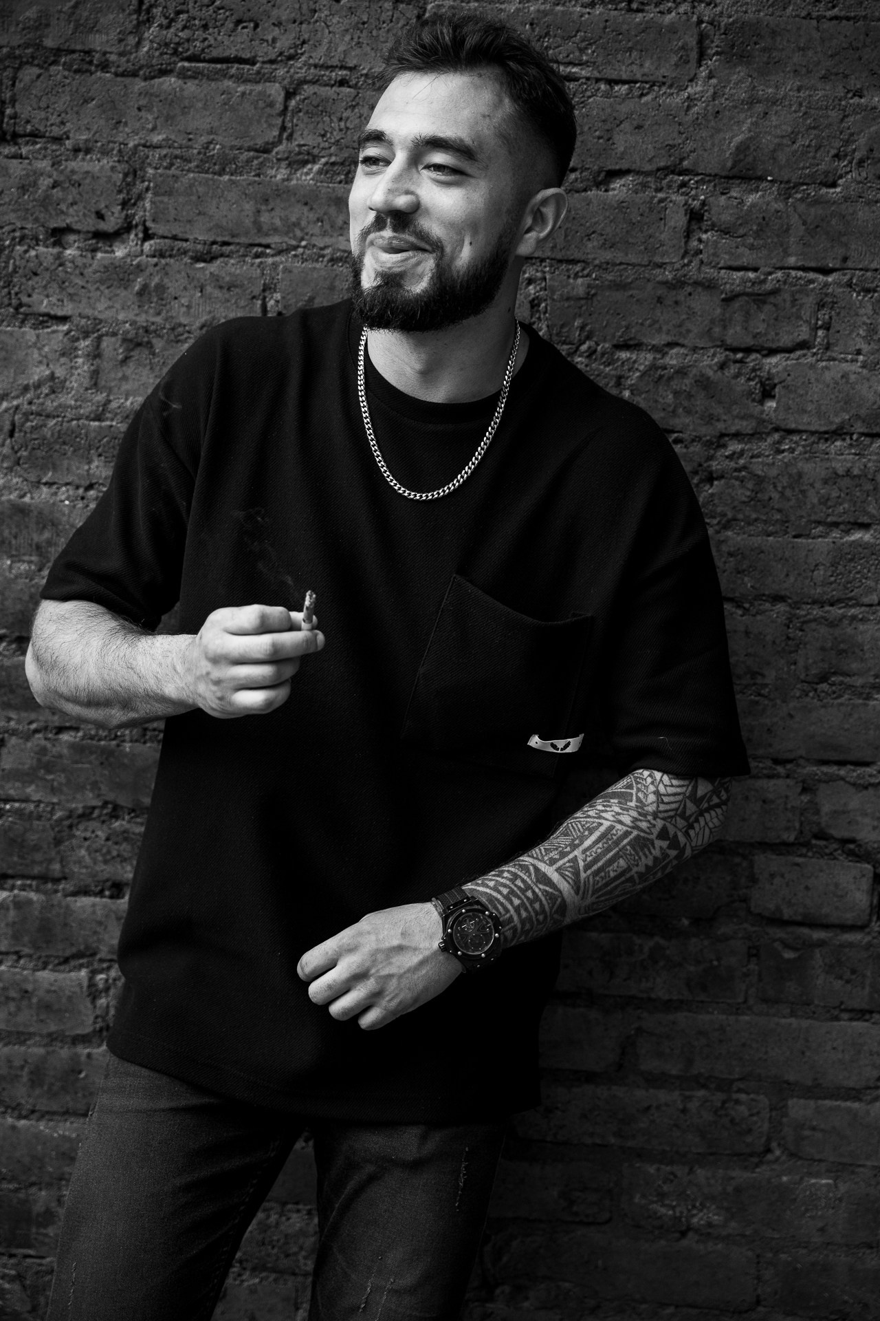 Мужчина с бородой в черной футболке курит сигарету на фоне кирпичной стены