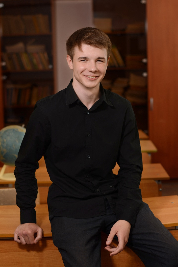 Парень-стиляга в чёрной рубашке сидя  на парте.