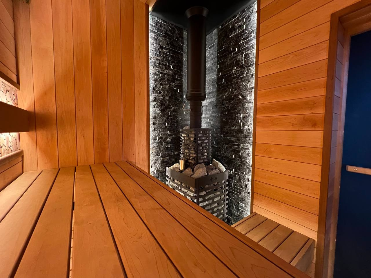 Семейная баня в Рощино из ольхи с можжевеловым панно, яркой печной зоной, закарнизной подсветкой и печью от производителя Aston. SaunaTopMasters