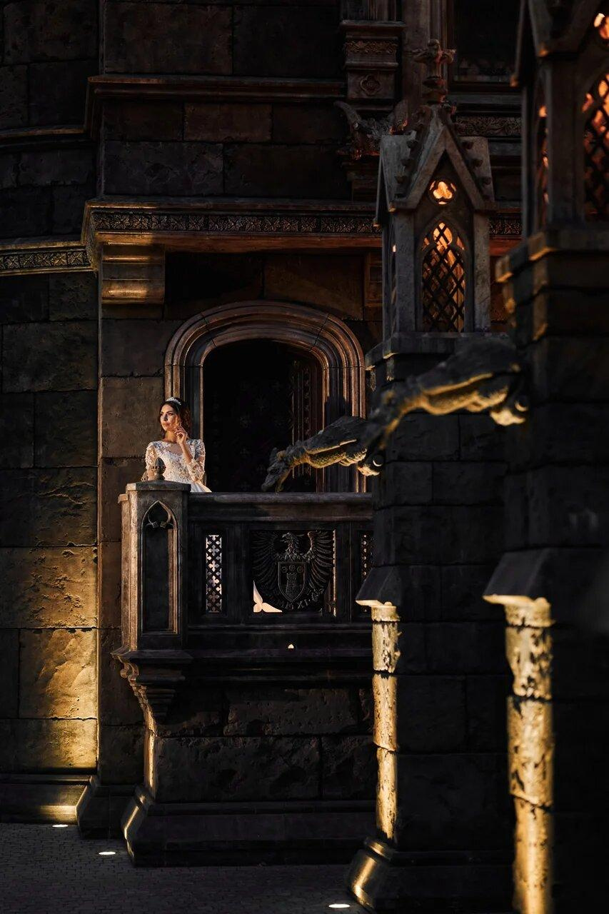 Женщина в тёмном наряде на балконе замка Гарибальди при вечернем освещении, подчёркивающем готический стиль архитектуры.