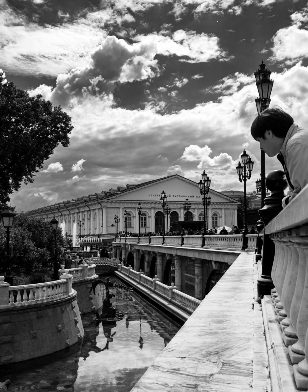 Черно-белая городская фотография: вид на канал с белым классическим зданием на противоположном берегу, отражение здания и неба в спокойной воде канала, фигура человека на парапете справа смотрит вниз, облачное небо, уличная фотография Москвы, Вадим Топорёнок, проект Манифест теней, городские отражения, геометрия воды, классическая архитектура, монохромная уличная съёмка.