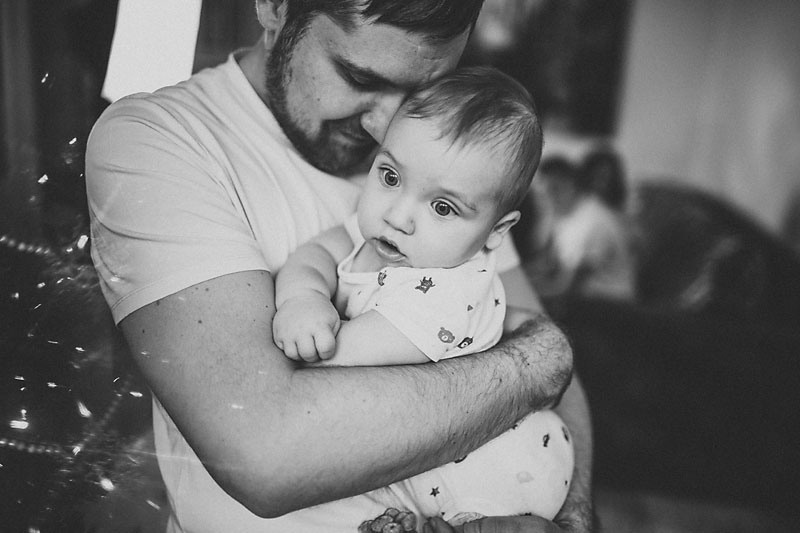 папа в белой майке и джинсах нежно обнимает своего новорожденного сына