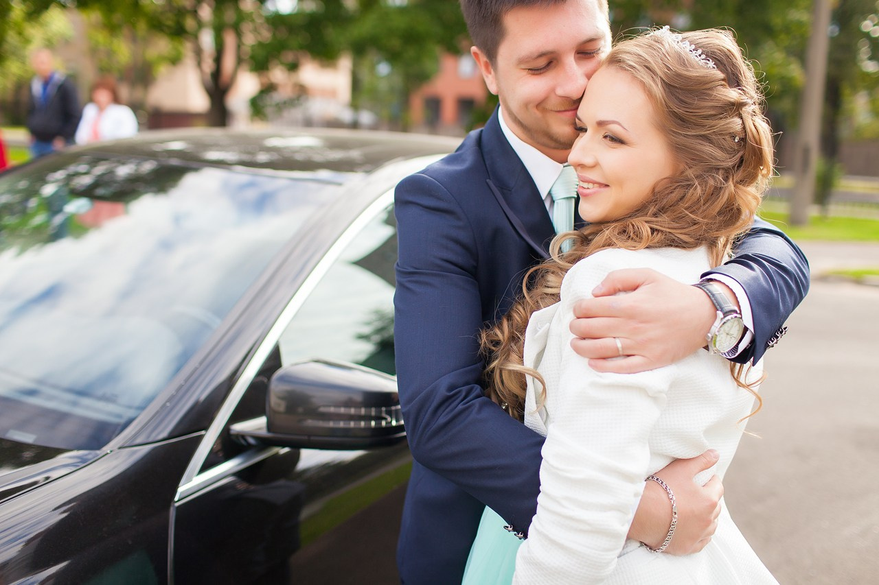 жених в синем костюме обнимает молодую невесту в диадеме на фоне автомобиля премиум классаж