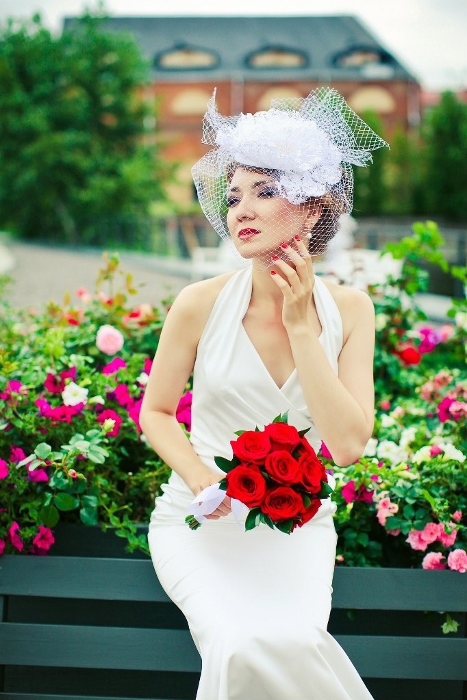 стильная невеста в ретро шляпке с вуалью держит в руке красный букет на фоне цветочной клумбы