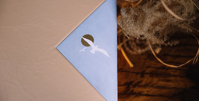 Макросъемка угла фотокниги. Тиснение белой чайки и золотого солнца на голубом бархатистом материале. Стык двух фактур обложки.