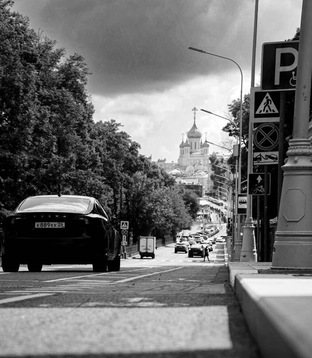 Черно-белая уличная фотография: городской проспект с деревьями по обеим сторонам, старинный автомобиль слева, дорожные знаки парковки и пешеходного перехода справа, в перспективе улицы видна сталинская высотка со шпилем, драматичное облачное небо, уличная фотография Москвы, Вадим Топорёнок, проект Манифест теней, архитектура сталинского ампира, городская перспектива, семи сестер, геометрия города, археология душ.