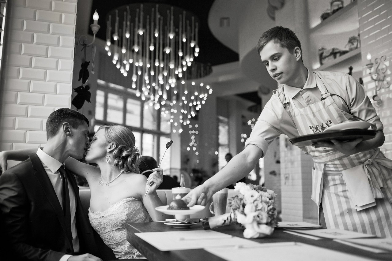 официант шикарного ресторана ставит на стол десерт пока молодожены целуются 