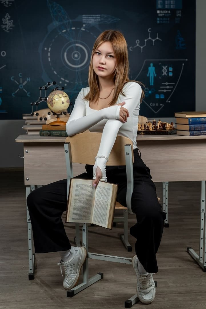 Девушка с учебников  в руках, сидит в классе оставив рабочее место позади.