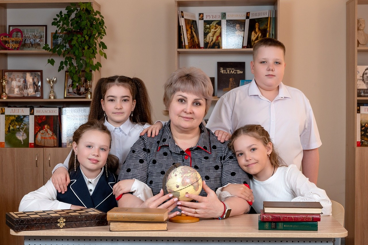 Ребята облепили учительницу сидя за партой в своём классе.