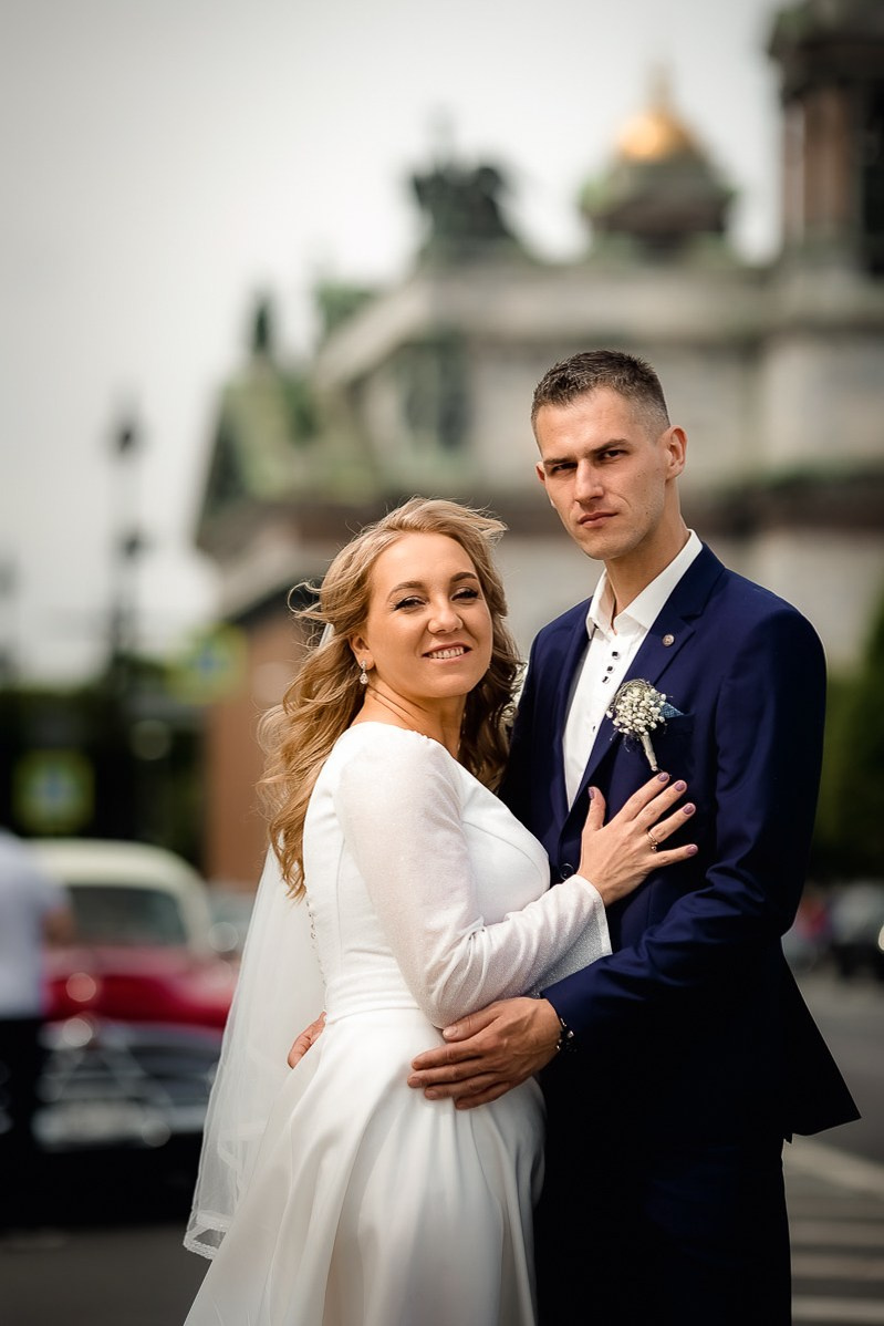 Жених и невеста смотрят в кадр на фоне Исаакиевского собора в Санкт-Петербурге. Вертикальный поясной кадр.