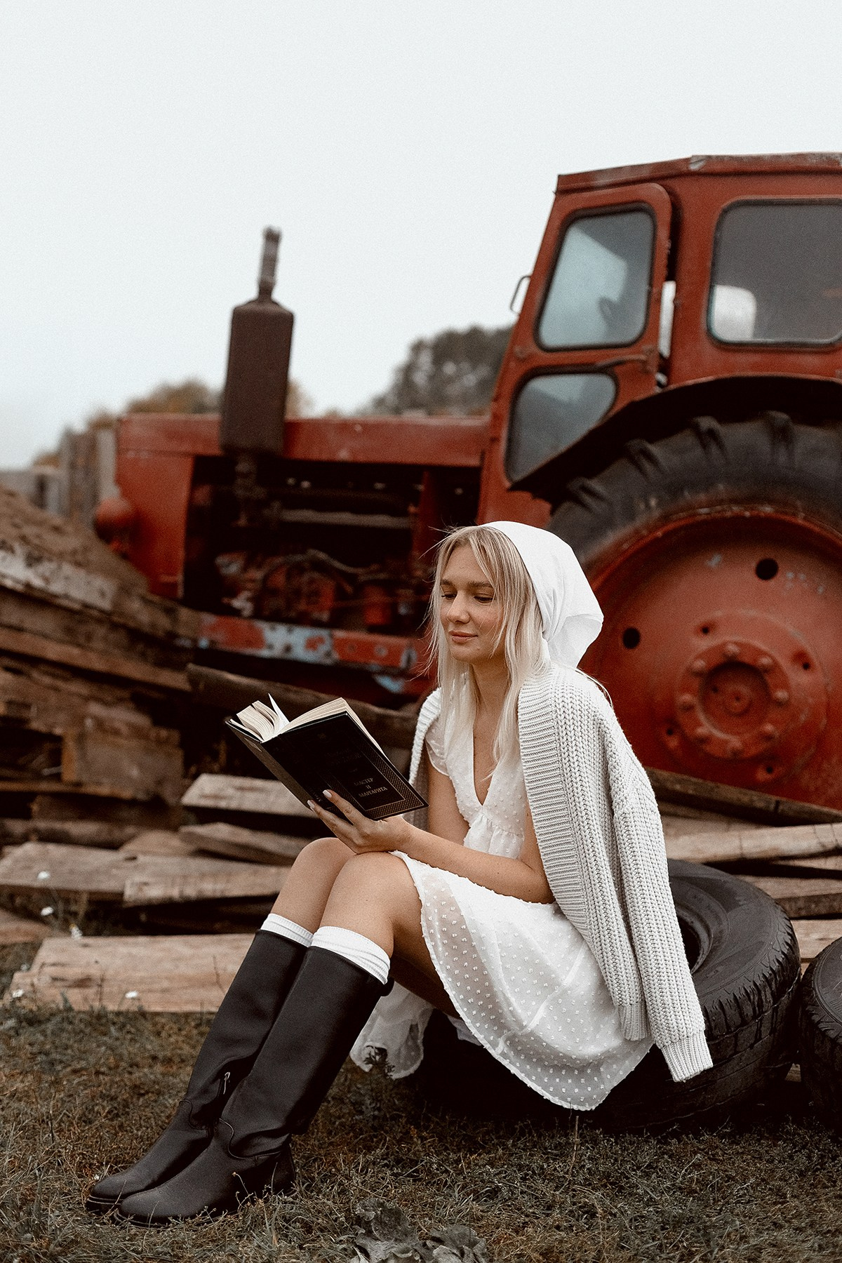 Одинокая девушка задумчиво сидит на автомобильной шине, а на фоне красный трактор. 