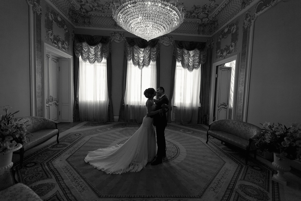 невеста в длинном свадебном платье целуется с женихом под хрустальной люстрой в загсе