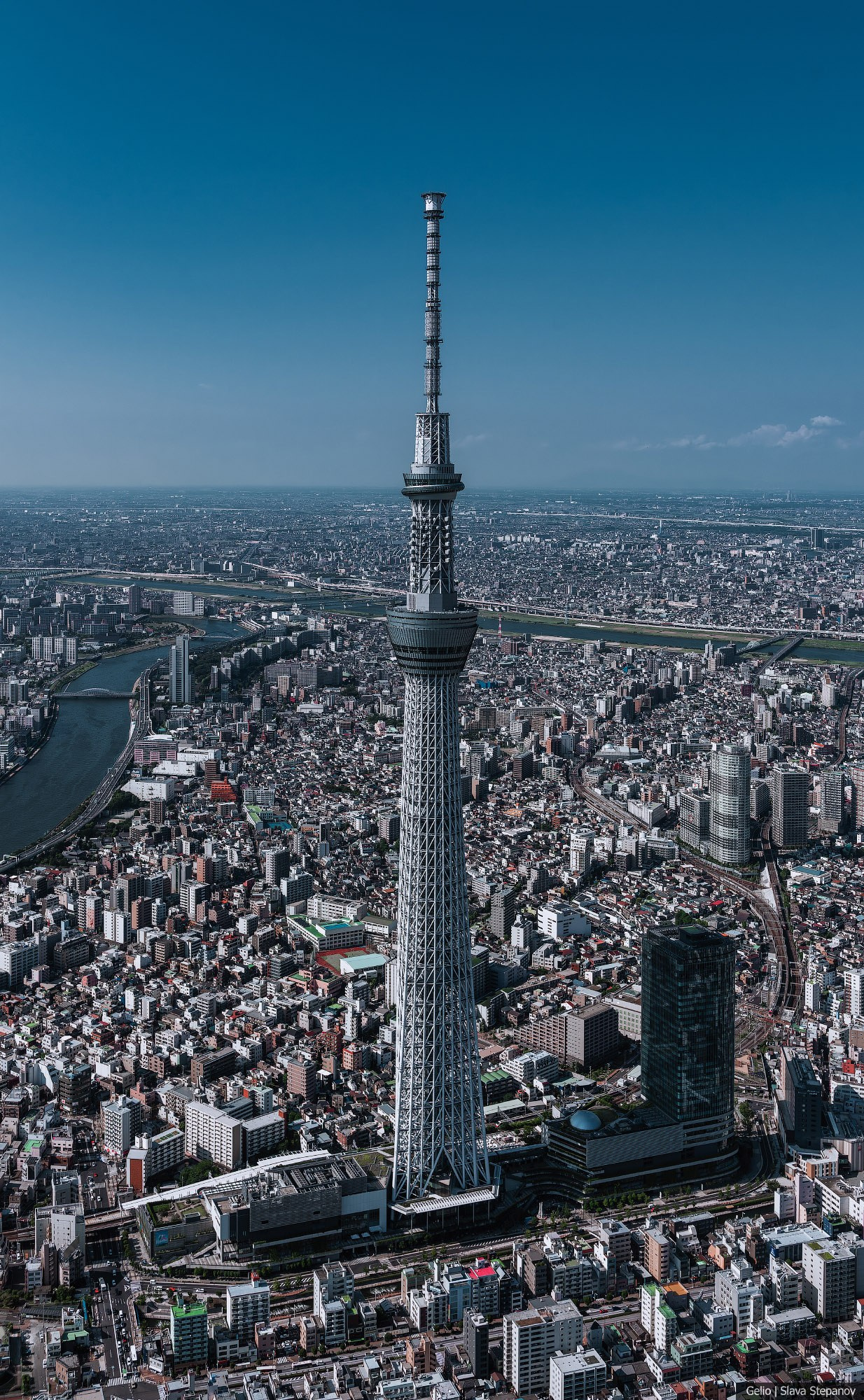 Аэрофотосъемка телевизионной башни Tokyo Skytree в Токио, Япония. Высотная конструкция расположена в центре кадра на фоне плотной городской застройки и русла реки под ясным синим небом. Автор фото: Слава Степанов.
