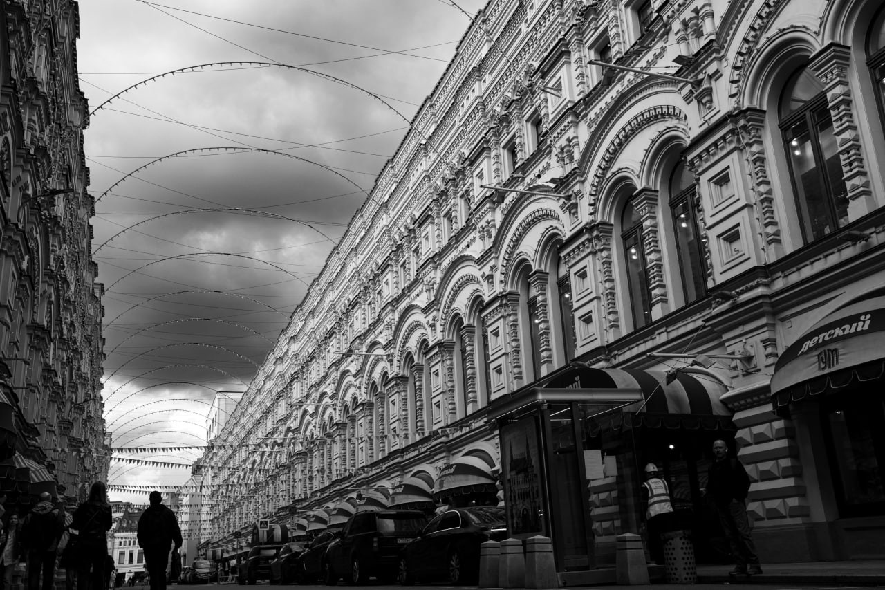 Черно-белая уличная фотография с широкоугольным объективом: длинный изогнутый фасад исторического здания с ритмичными арочными окнами, над улицей натянуты дугообразные провода, внизу силуэты прохожих, драматичное облачное небо, искажённая перспектива, городская архитектура Москвы, уличная фотография, Вадим Топорёнок, проект Манифест теней, геометрия города, городские текстуры, монохромная съёмка.