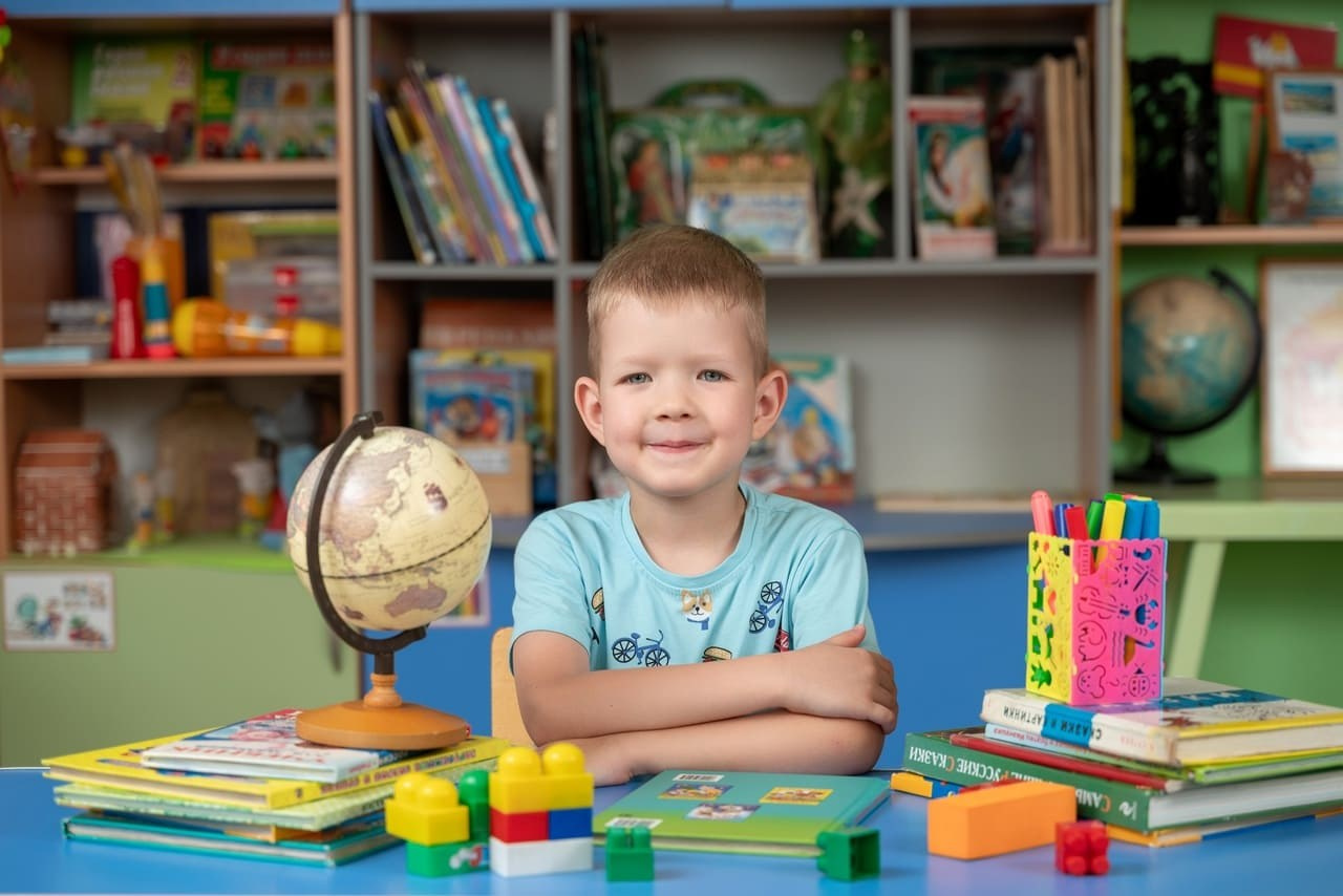 Мальчик за партой в голубой футболке мило сидит перед фотографом.