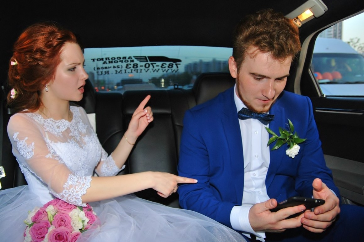 Свадебная фотосъёмка, молодожены едут в автомобиле, жених сидит в телефоне, недовольная невеста грозит жениху пальцем