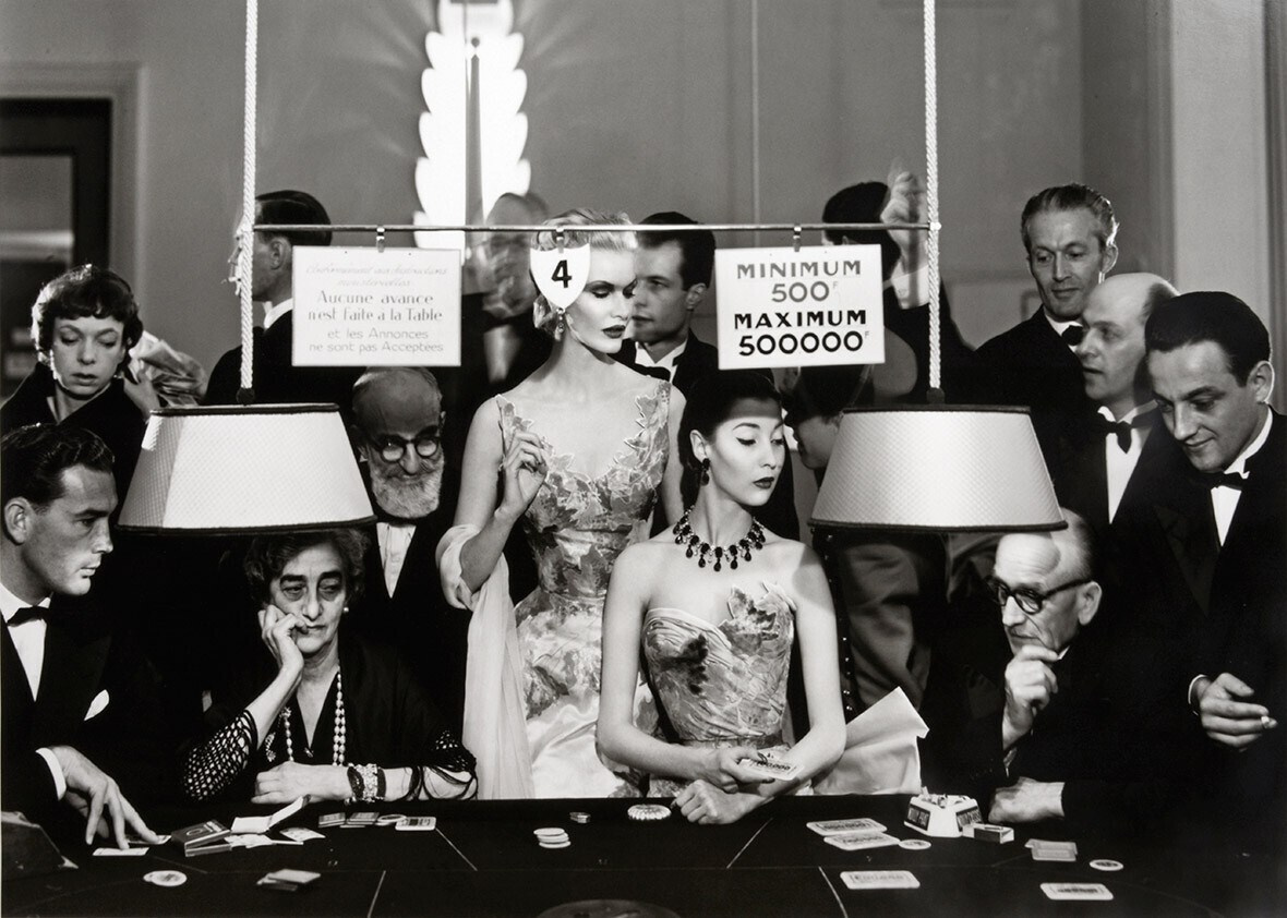 Санни Харнетт и Алла. Вечерние платья от Balmain, Casino Le Touquet, август 1954. Санни Харнетт и Алла здесь — не просто «лица моды», а героини почти кинематографической сцены: вокруг них — публика у игрового стола, лампы, таблички с лимитами ставок и надпись на французском о запрете авансов и объявлений. Такой приём — встраивать высокую моду в реалистичный, почти репортажный контекст — был фирменным ходом Аведона: он ломал студийную условность и делал фэшн‑кадр живым. Платья Balmain подчёркивают эстетику 1950‑х: скульптурные корсетные силуэты, фактурные ткани, деликатная отделка — всё это работает на образ «роскоши без пафоса». При этом позы моделей и их выражения лиц намеренно сдержанны: Аведон избегал «улыбок по команде», предпочитая психологическую глубину и даже лёгкую отстранённость. 