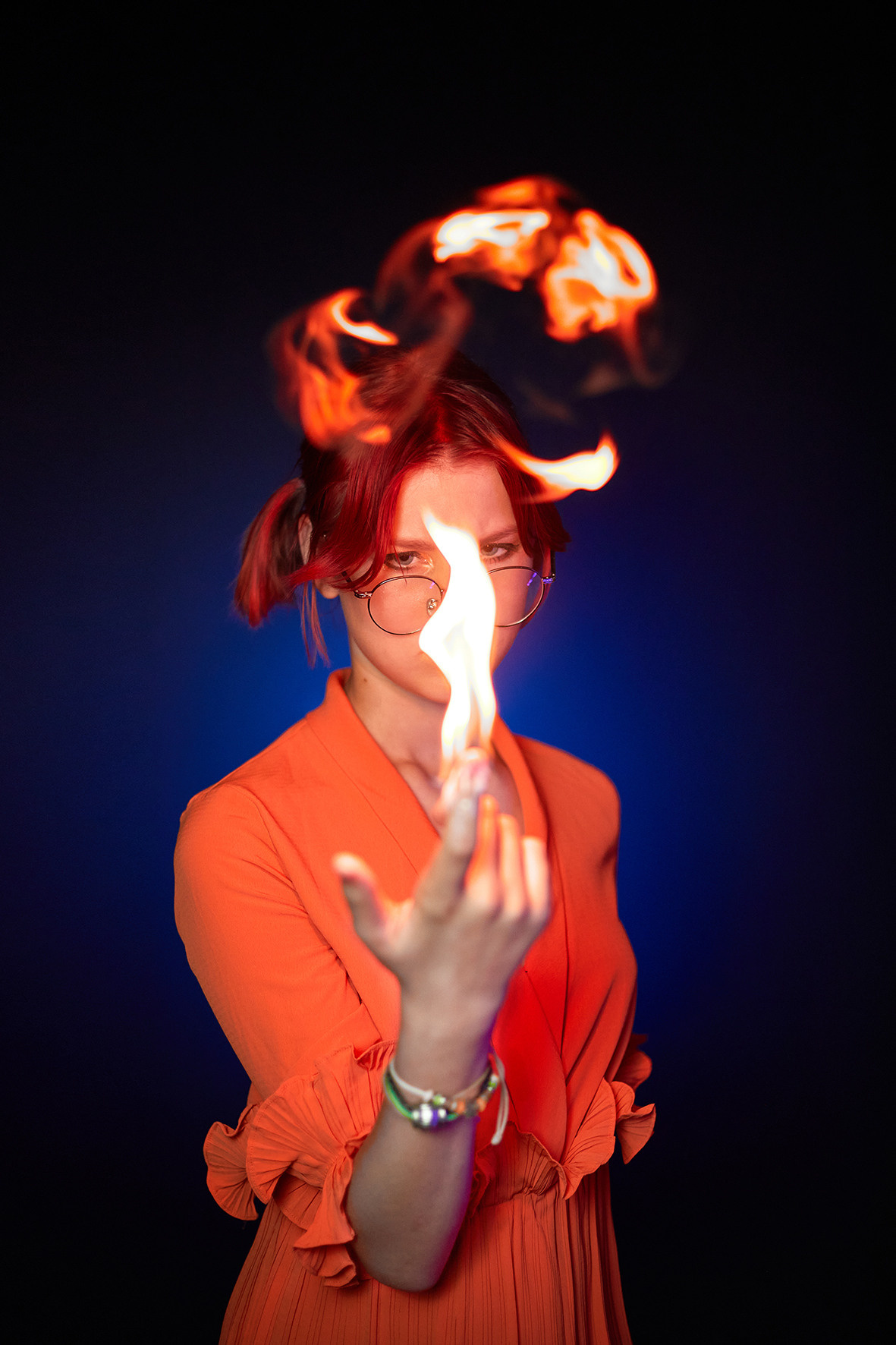 Студийный портрет девочки в очках с красными волосами в оранжевом платье на синем фоне. Девочка с красными волосами держит в руках огонь. Фотограф. Тимашевск. Кубань. Краснодарский край.