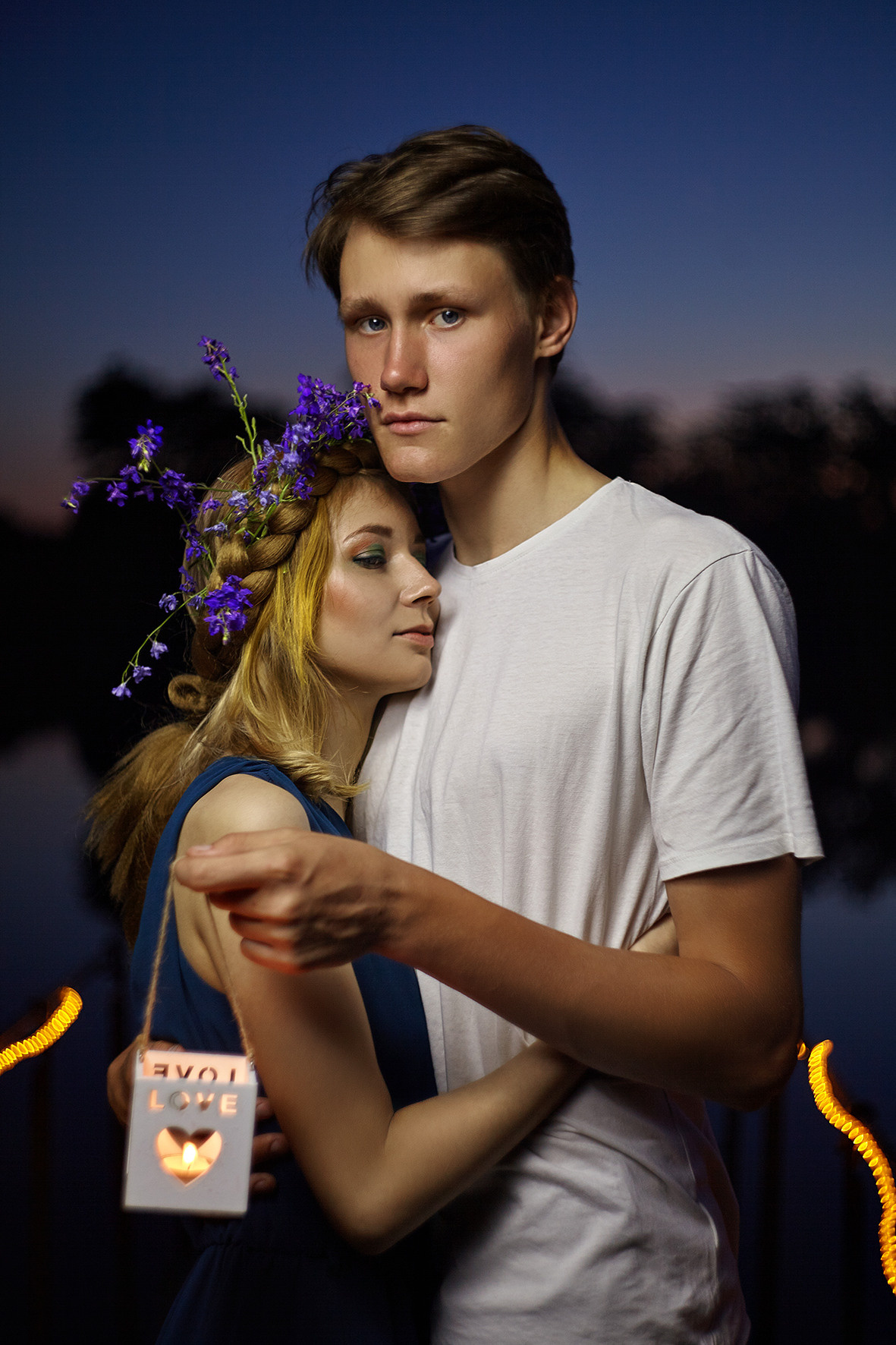 Влюбленная пара на мосту над рекой в часы заката. Парень обнимает девушку с цветочным венком на голове. Влюбленные держат в руках декоративную свечу в подсвечнике с сердечком. Фотограф. Тимашевск. Кубань. Краснодарский край.