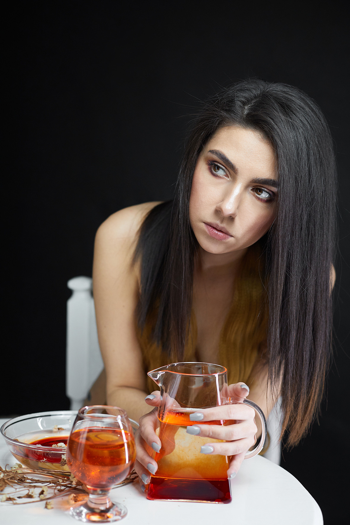 Студийный портрет обнаженной девушки с графином красной воды на черном фоне.  Обнаженная девушка с длинными волосами сидит за белым столом с прозрачной посудой и окровавленной водой и сухоцветами. Фотограф. Тимашевск. Кубань. Краснодарский край.