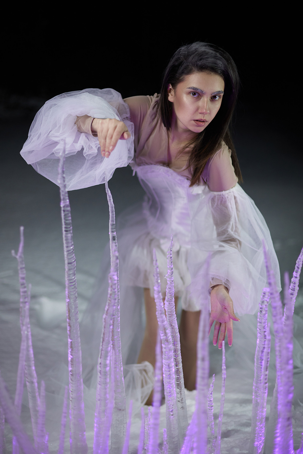 Девушка в белом платье с рукавами-воланами стоит в снегу за изгородью из замерзших сосулек. Сосульки торчат из снега. Снежная королева. Фотограф. Тимашевск. Кубань. Краснодарский край.