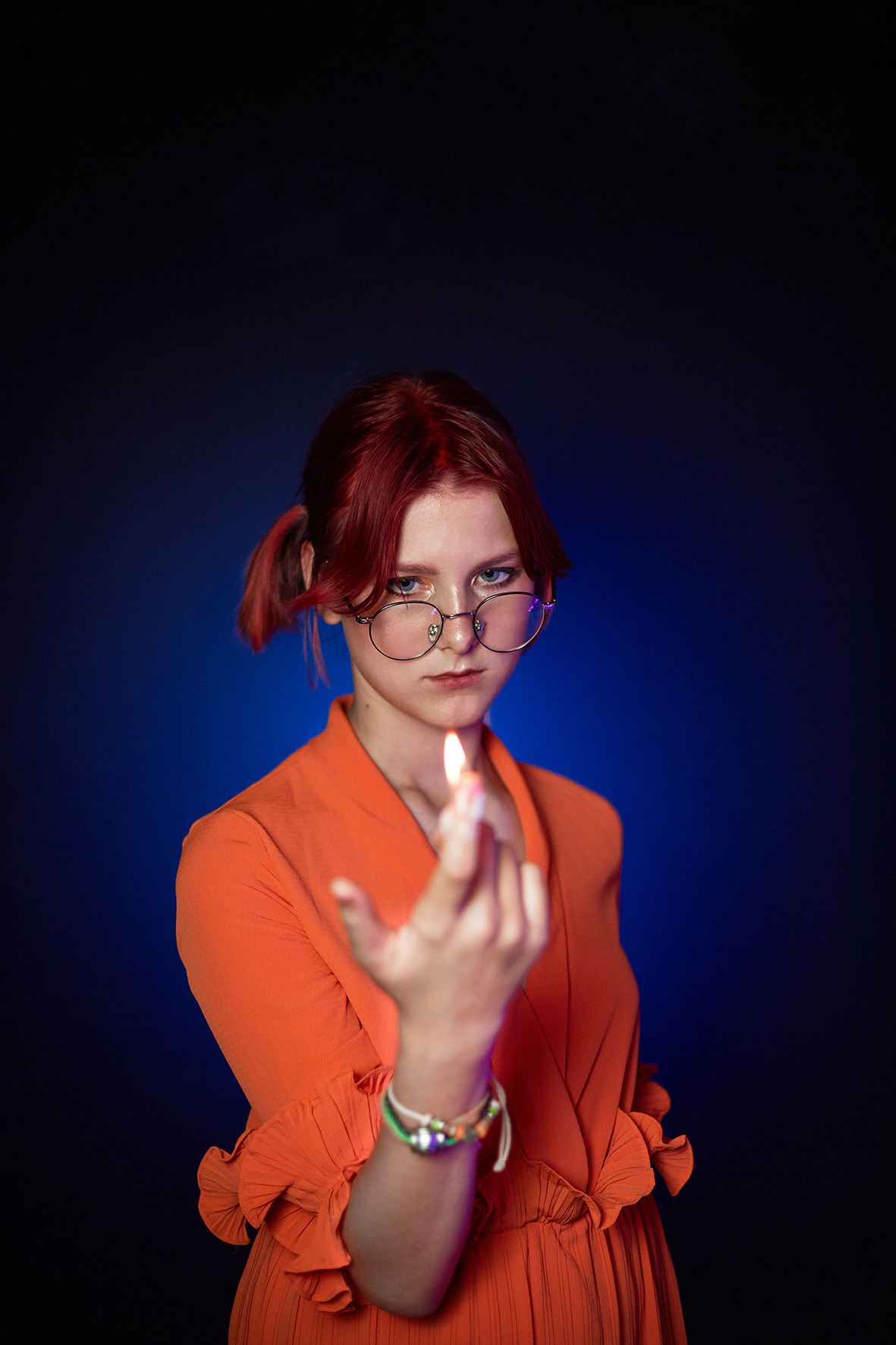 Студийный портрет девочки в очках с красными волосами в оранжевом платье на синем фоне. Девочка с красными волосами держит в руках огонь. Фотограф. Тимашевск. Кубань. Краснодарский край.