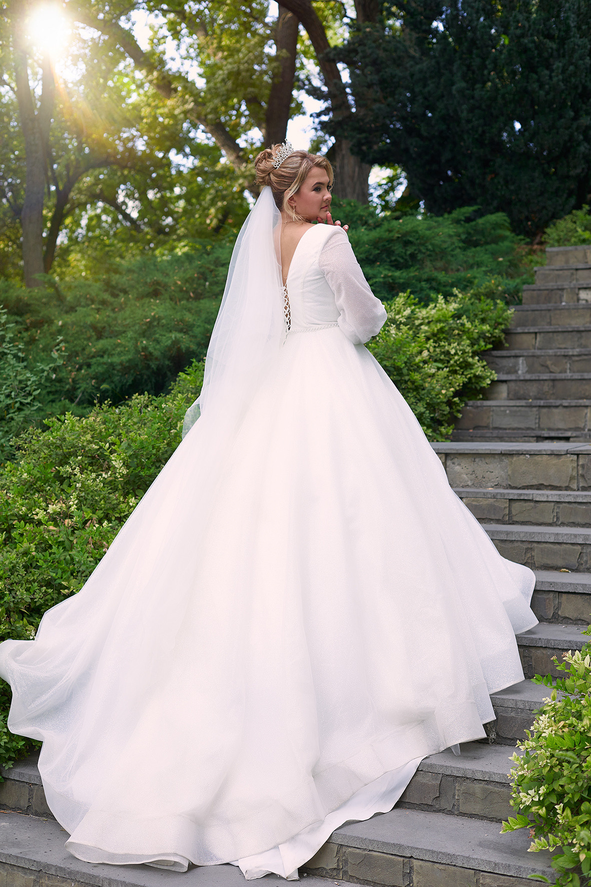 Невеста в платье со шлейфом стоит на ступенях в зеленом парке в часы заката. Фотограф. Тимашевск. Кубань. Краснодарский край.