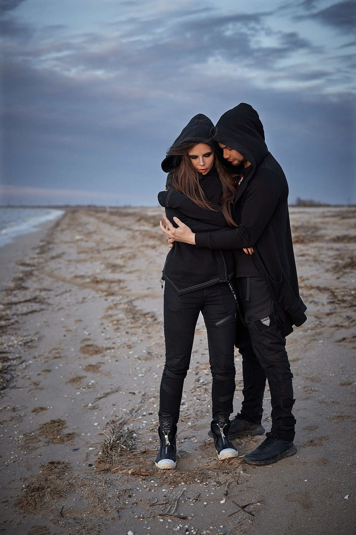 Лавстори на берегу моря во время заката. Море зимой. Песок под ногами. Линия берега и волны. Парень и девушка в черной одежде обнимаются на берегу моря на закате. Приморско-Ахтарск. Фотограф. Тимашевск. Кубань. Краснодарский край.