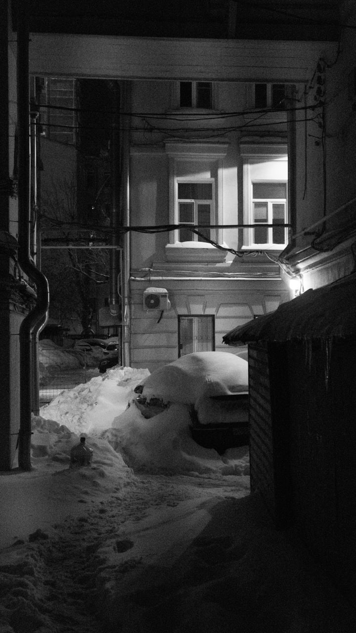 Черно-белая ночная уличная фотография: заснеженный городской двор с освещенными окнами жилого дома, массивные сугробы на переднем плане, водосточные трубы и провода на фасаде, резкий контраст теплого внутреннего света и холодной внешней темноты, атмосфера зимнего затишья, уличная фотография Москвы, Вадим Топорёнок, проект Манифест теней, городская геометрия, свет и тень в урбанистике, археология душ, монохромная зимняя сцена.