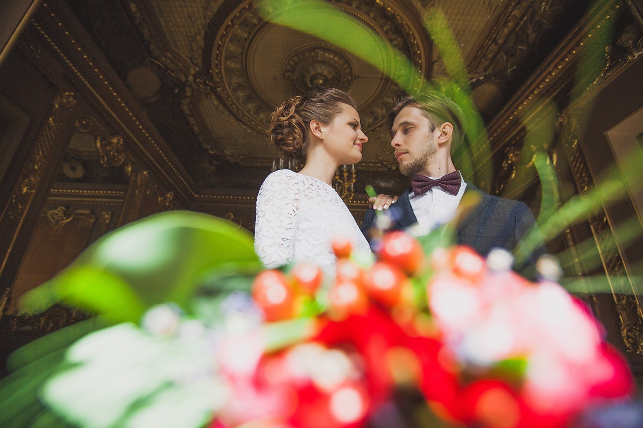 жених и невеста в парадном зале дворца с большим красным букетом на переднем плане