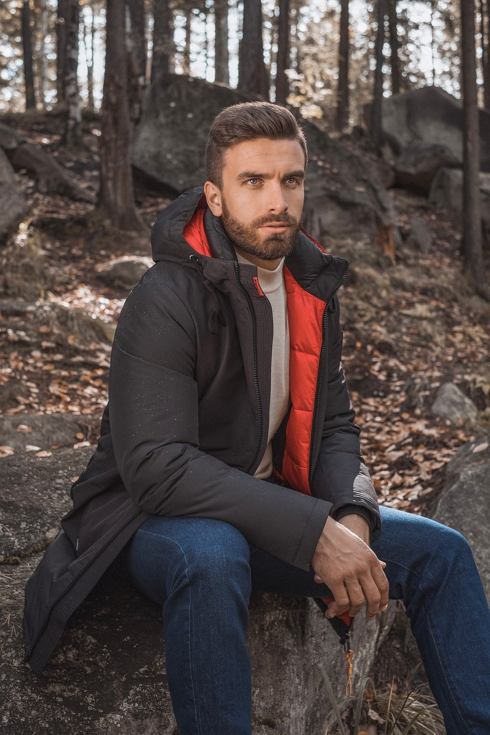Мужчина с бородой в черной теплой куртке с капюшоном и синих джинсах сидит на камне в лесу