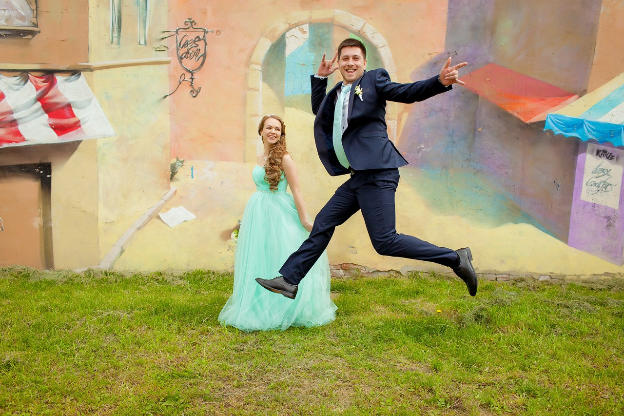 невеста в платье цвета тиффани позирует на фоне прыгающего жениха у стены с граффити 