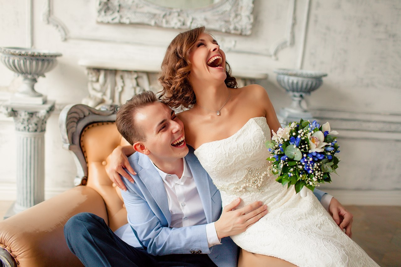 жених в голубом пиджаке и невеста в роскошном платье с жемчугом смеются в старинном кресле