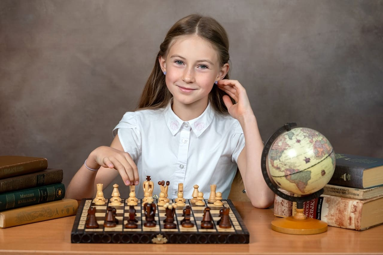 Девочка фотографируется за игрой в шахматы на коричневом фоне.