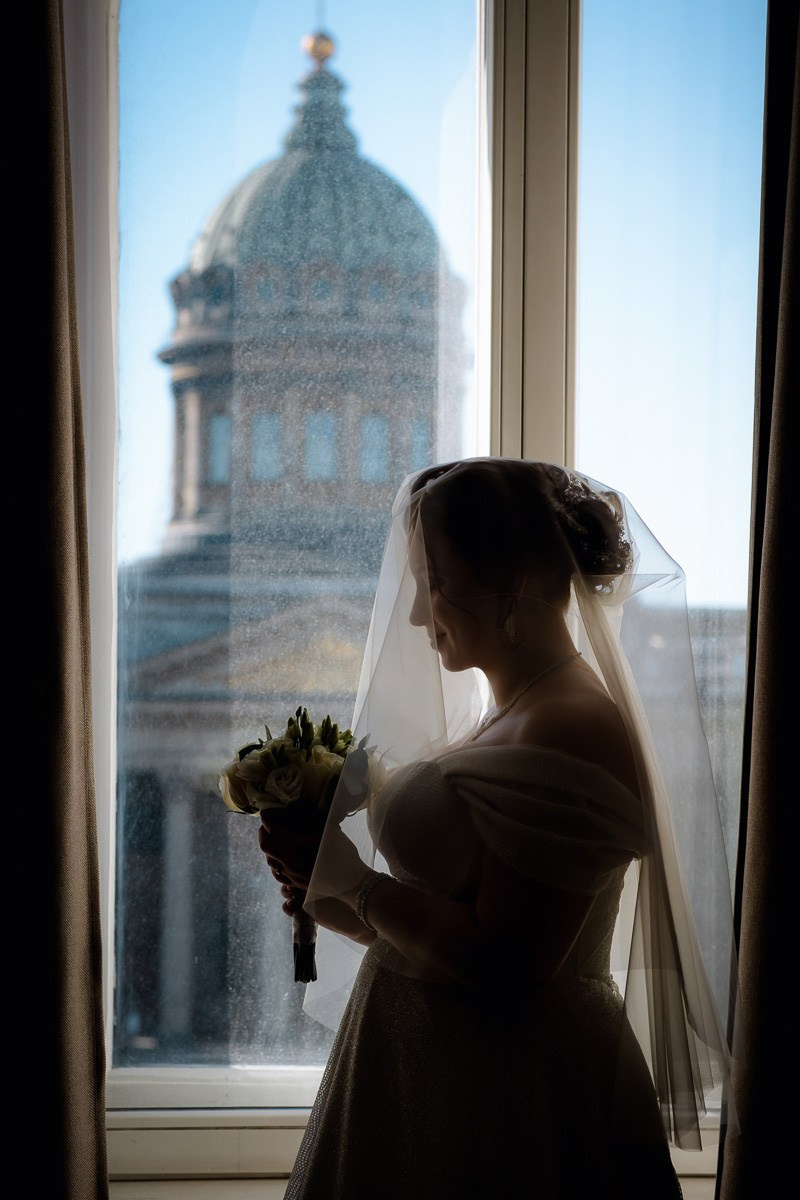 Невеста в отеле смотрит на букет , а за окном Казанский собор Петербурга