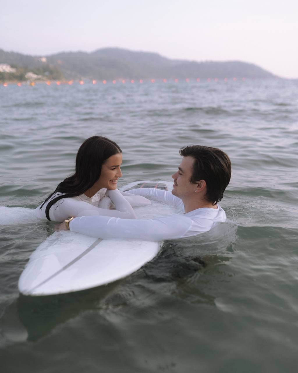 Пара обнимается в море рядом с доской — тёплый кадр