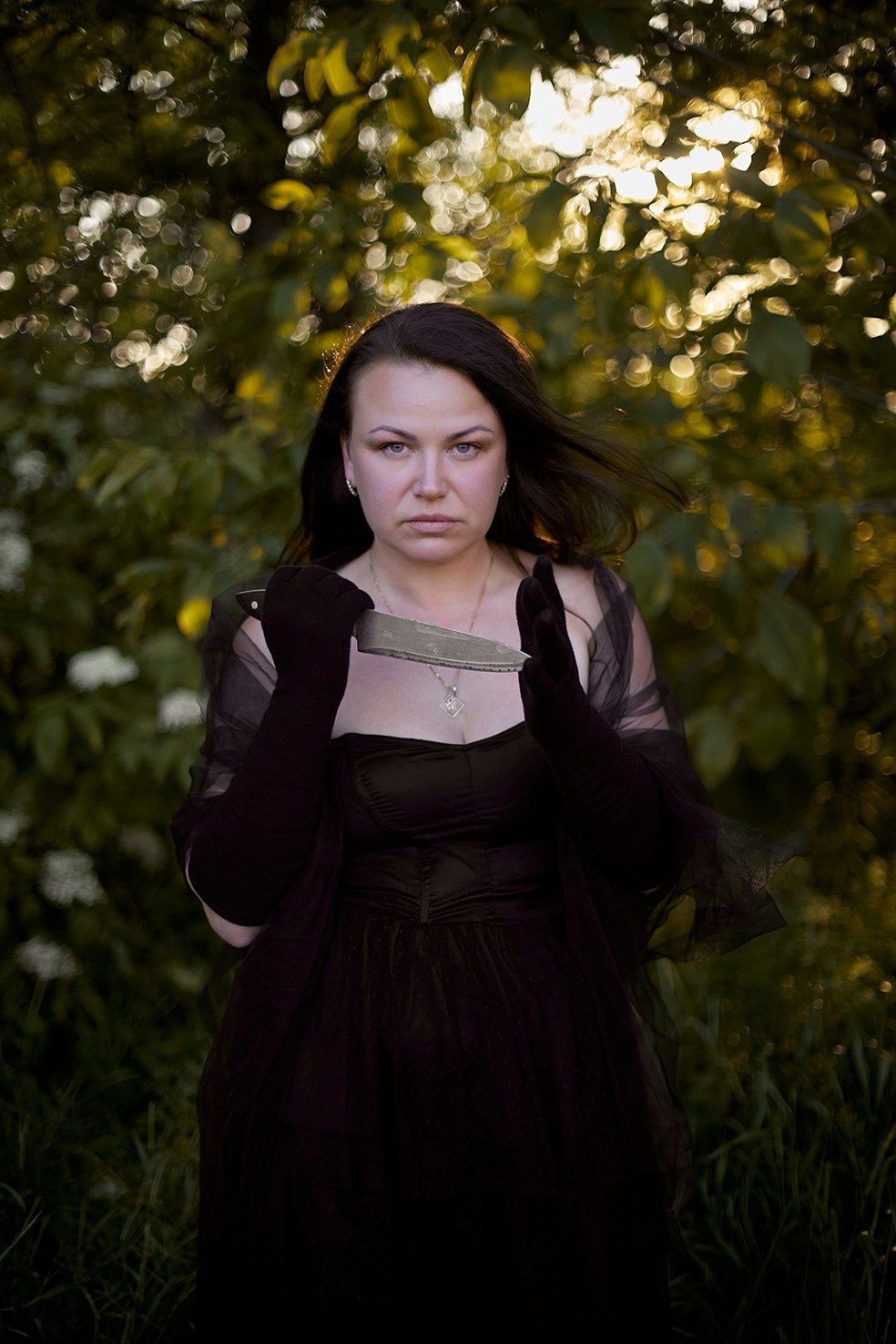 Фотограф в Москве. Креативное фото. Девушка в черном платье и с ножом стоит в лесу. Закат. 