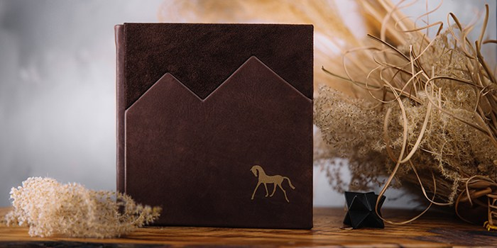Фотокнига ручной работы в коричневой кожаной обложке с дизайном в виде горных пиков. Сочетание гладкой кожи и ворсистой замши, украшенное золотистым тиснением силуэта лошади.