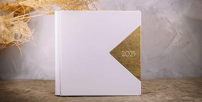 Белая минималистичная фотокнига с золотым треугольником и надписью 2025. Книга на фоне натуральных текстур и колосьев.