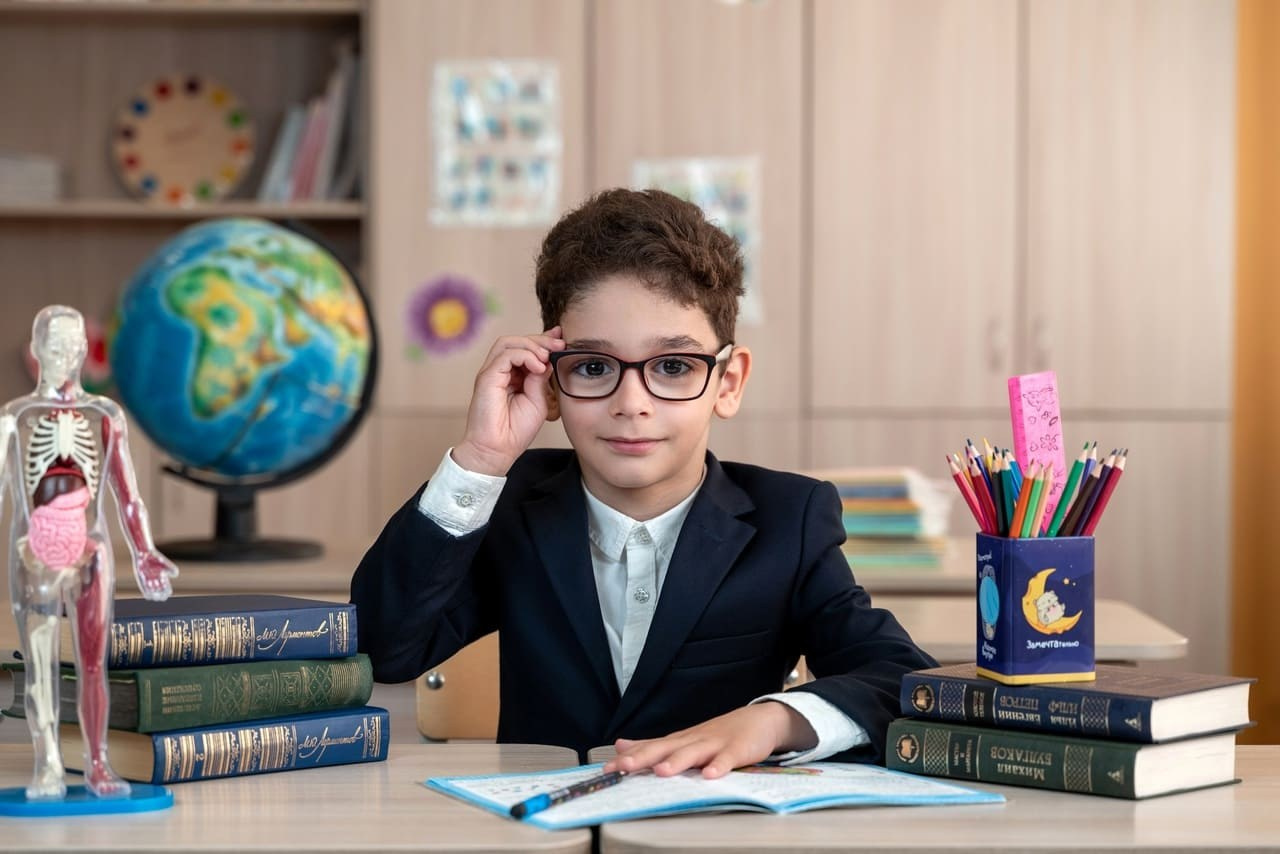 Мальчик в черном пиджаке стильно теребит очки на фотокамеру.