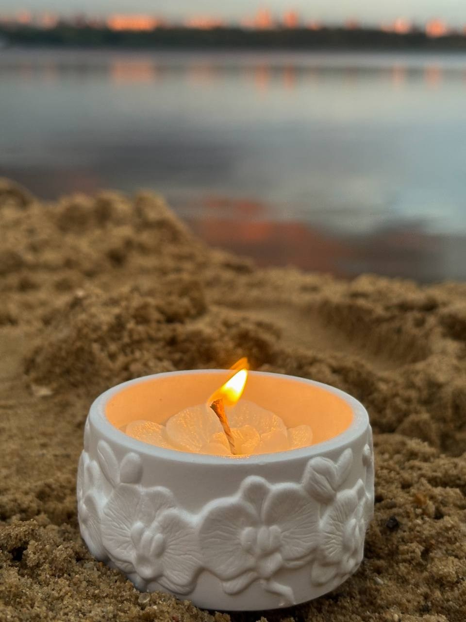 Фото белой свечи в керамическом стакане на песке у воды на закате.