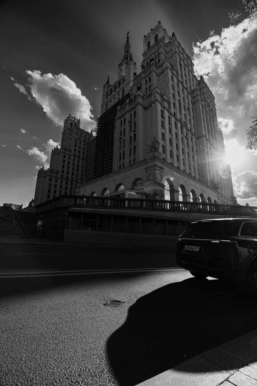 Черно-белая городская фотография: монументальное сталинское высотное здание, снятое с нижнего ракурса, острый шпиль пронзает драматичное облачное небо, яркий солнечный контровой свет создает силуэтный контраст, на переднем плане темный автомобиль отбрасывает длинную резкую тень на асфальт, уличная фотография Москвы, Вадим Топорёнок, проект Манифест теней, геометрия власти, архитектурный монументализм, игра света и тени, широкоугольная перспектива, городская археология.
