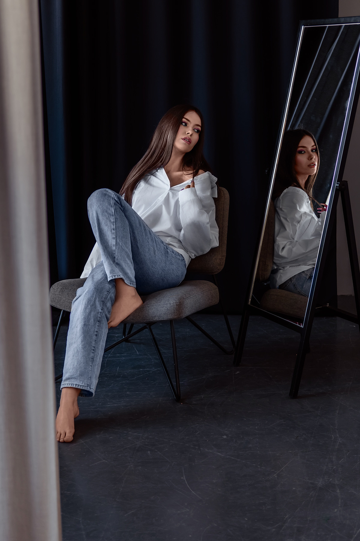 Девушка сидит на стуле в фотостудии и смотрит на свое отражение в зеркале.
