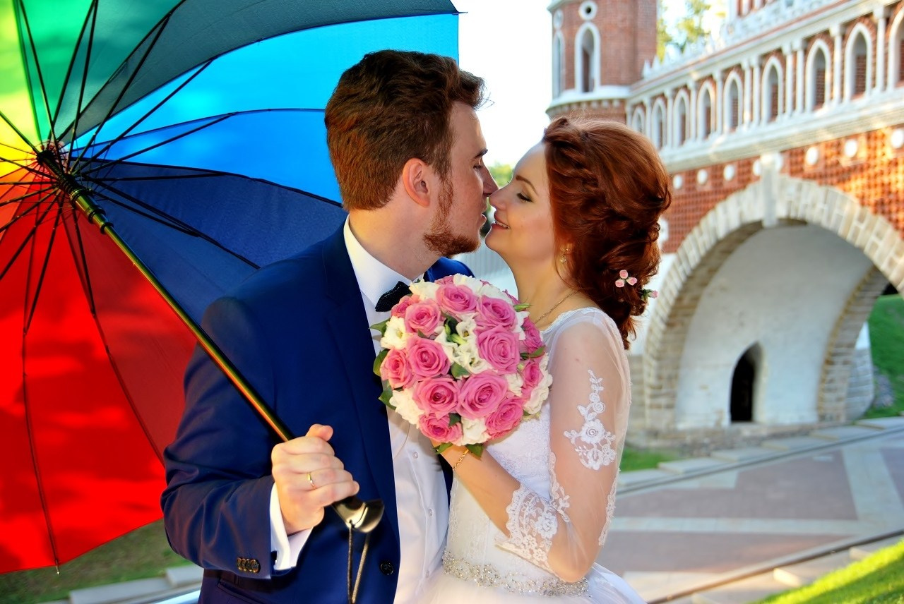 Свадебная фотосъёмка в Царицыно летом на закате, поцелуй новобрачных на фоне каменного моста из красного и белого камня и арки, жених держит в руке яркий разноцветный зонт, невеста держит в руке бело-розовый букет