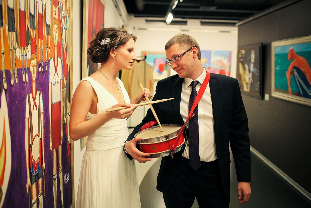 невеста с барабанными палочками и жених с барабаном смотрят друг другу в глаза в музее