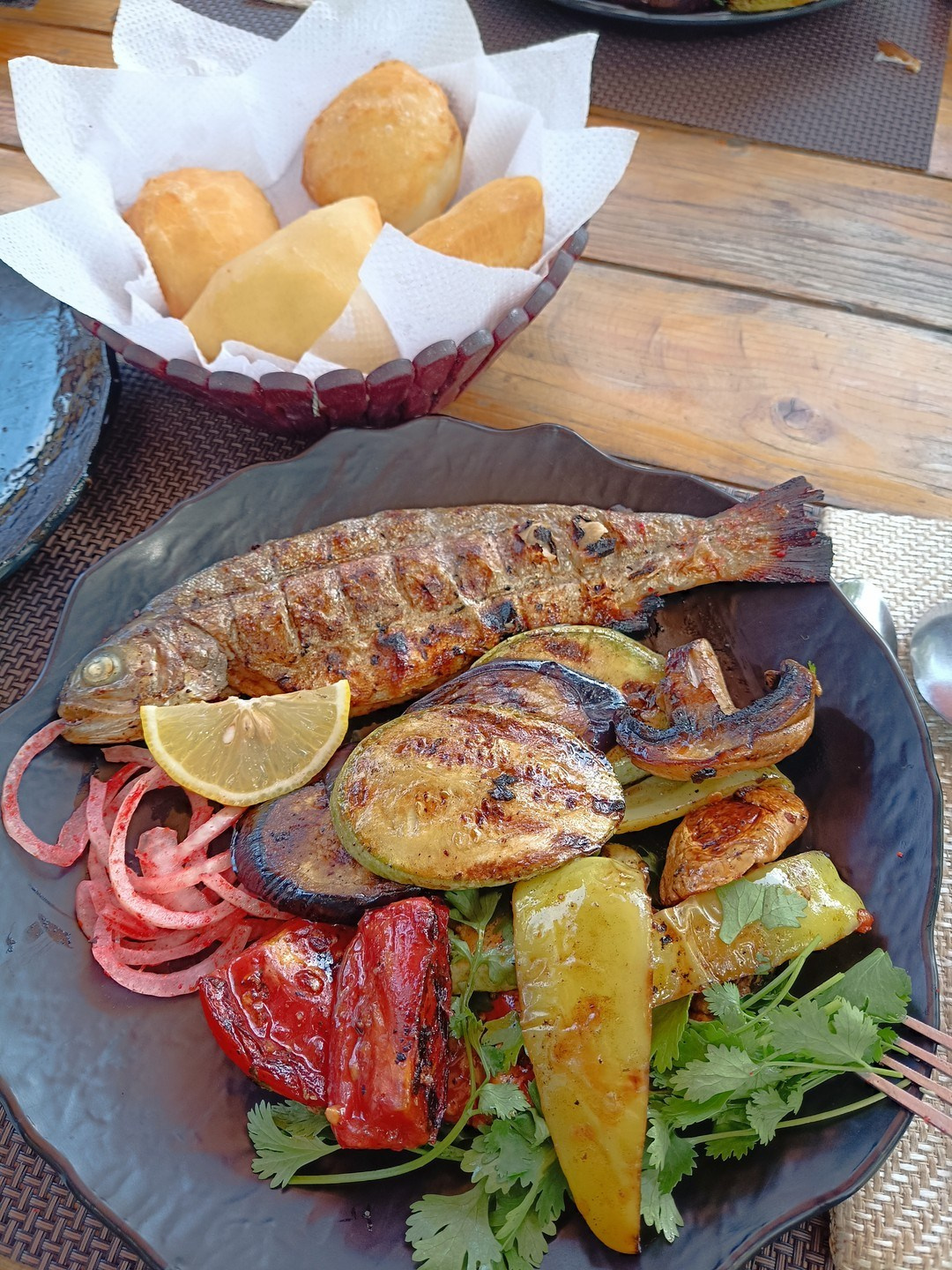 Аппетитное блюдо с запеченной рыбой, гарниром из запеченных овощей (баклажаны, перец, картофель) и свежей зеленью. Рыба украшена долькой лимона и тонкими ломтиками маринованного лука. На заднем плане в корзине лежат небольшие румяные пирожки на бумажной салфетке. Всё это расположено на деревянном столе, создавая атмосферу уютного и вкусного обеда на свежем воздухе в походе на водопад Медвежий.