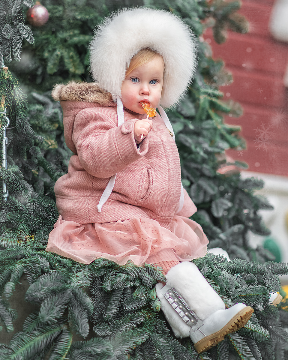 Ребенок сидит возле ёлки с леденцом во рту.