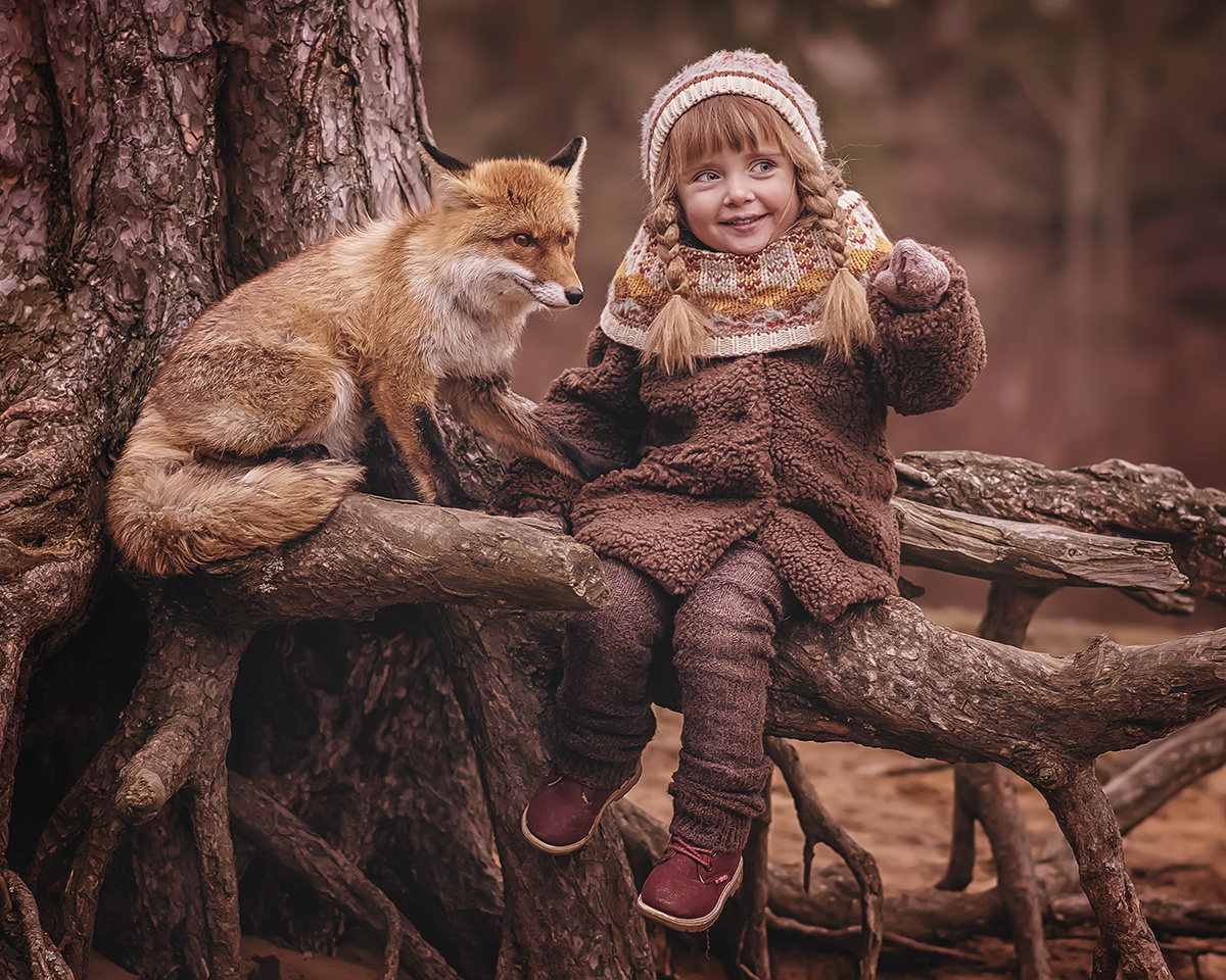 Девочка сидит на корнях большого дерева рядом с лисой и что-то ей показывает пальцем.