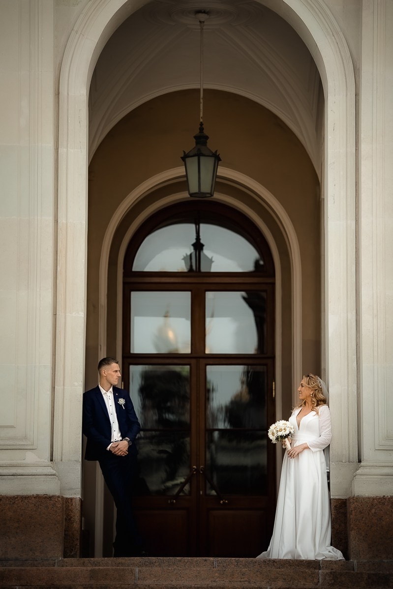 Жених и невеста смотрят в кадр, стоя в арке на крыльце заксобрания в Санкт-Петербурге. Вертикальный ростовой кадр.